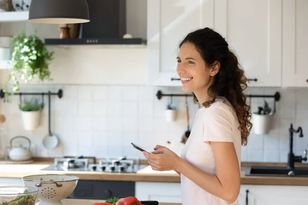 Joyeux femme attrayante en utilisant le téléphone, debout dans la cuisine moderne, cuisine — Photo