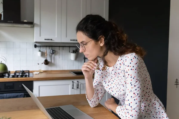 Mujer enfocada con gafas mirando la pantalla del portátil en la cocina — Foto de Stock