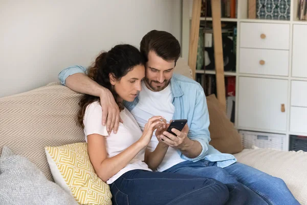 Giovani coppie che si abbracciano, usando lo smartphone insieme, rilassandosi sul divano accogliente — Foto Stock