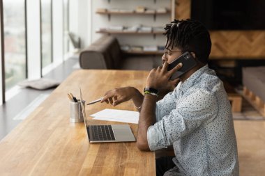 Afrikalı Amerikalı adam bilgisayarda çalışıyor ve cep telefonuyla konuşuyor.