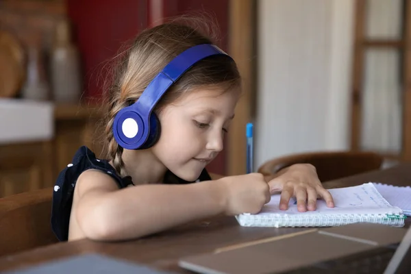 Nahaufnahme eines kleinen Mädchens mit Kopfhörern, das Notizen schreibt und online lernt — Stockfoto