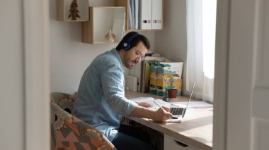 Kulaklık takan, internetten ders çalışan, notlar yazan, dizüstü bilgisayar kullanan bir adam.