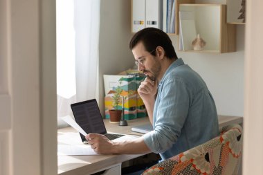 Gözlük takan, elinde belge tutan, okuyan, evde çalışan kendinden emin bir adam.