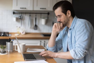 Düşünceli adam çeneye dokunurken dizüstü bilgisayar kullanıyor, sorunu çözüyor.