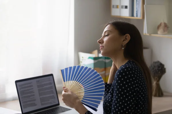 Close up exhausted woman waving paper fan at workplace