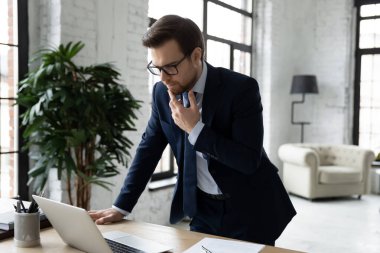 Düşünceli iş adamı, strateji düşünüyor, dizüstü bilgisayar kullanıyor, masanın yanında duruyor.