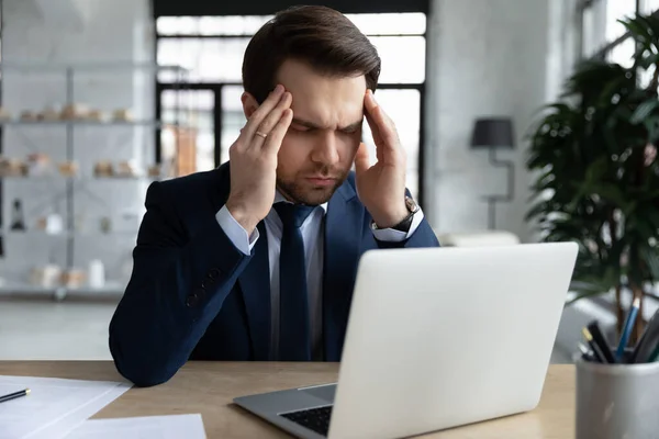 Cansado empresario estresado tocar templos, sentado en el escritorio con el ordenador portátil — Foto de Stock