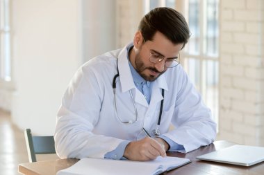 Erkek doktor masasına oturur ve tıp dergisine yazar.