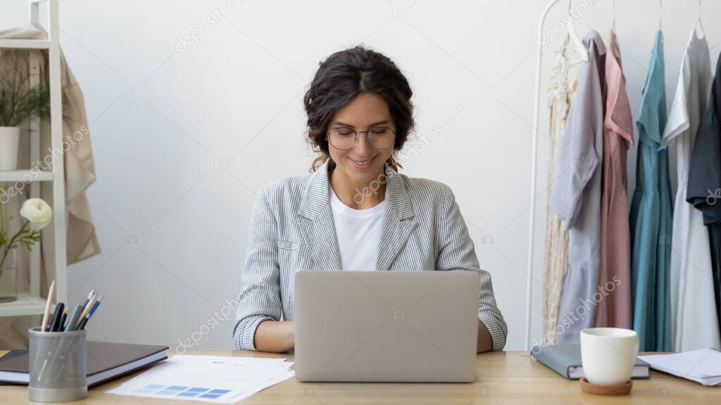 Happy young businesswoman self-employed tailor sitting at desk using laptop