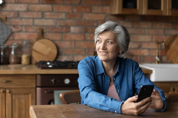Мечтательная седовласая женщина средних лет держит в руках смартфон. — стоковое фото