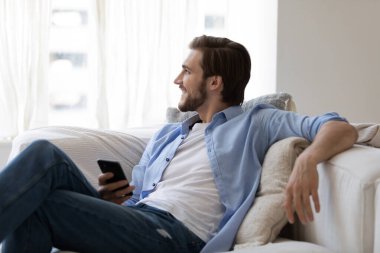 Mutlu genç adam kanepede dinleniyor, akıllı telefonu elinde tutuyor..