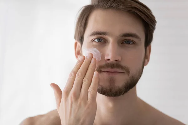 Happy young man applying moisturizing cream on cheek. — Stock Photo, Image