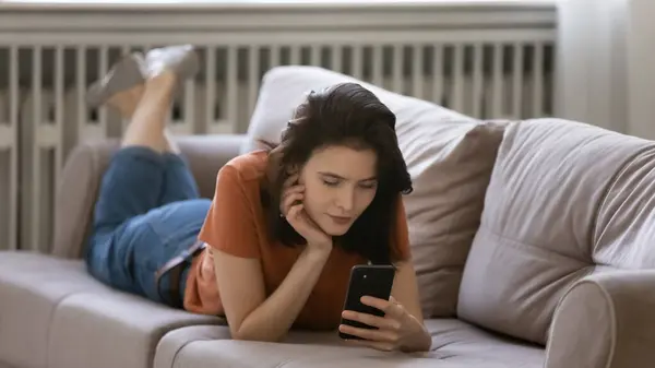 Concentrated millennial female lying on couch on belly using smartphone — Stock Photo, Image