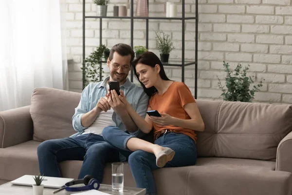 Jóvenes cónyuges felices sentados en el sofá usando teléfonos inteligentes compartiendo información — Foto de Stock
