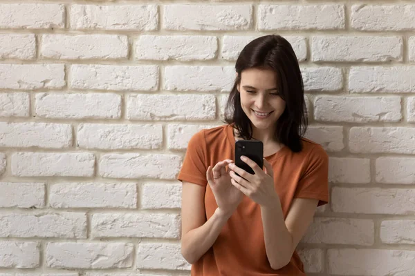 Glimlachende jongedame in de buurt van muur betrokken bij het gebruik van mobiele telefoon — Stockfoto