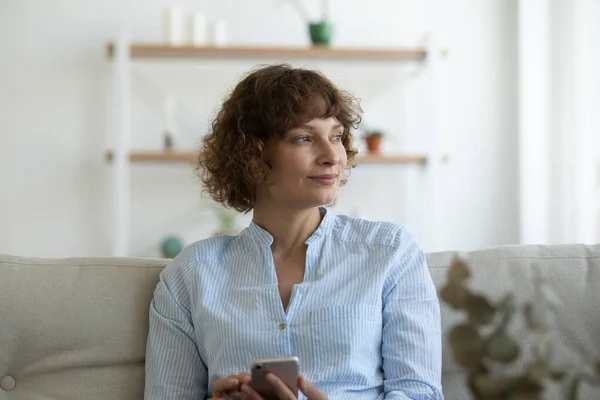 Lächelnde junge Frau sitzt auf Couch und hält Handy gedanklich in der Hand — Stockfoto