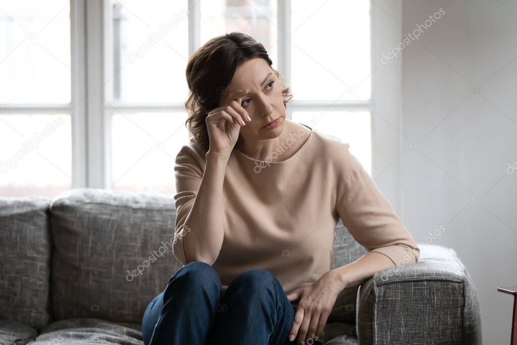 Thoughtful mature woman touching forehead, sitting on couch alone