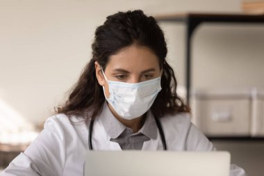 Yüz maskeli kadın doktor klinikteki bilgisayarda çalışıyor.