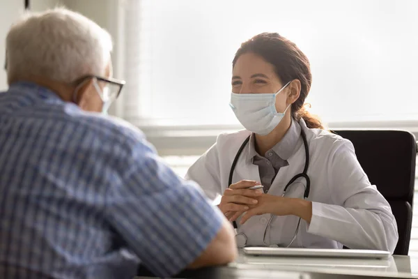 Yüz maskeli gülümseyen doktor son sınıf erkek hastaya danışıyor. — Stok fotoğraf