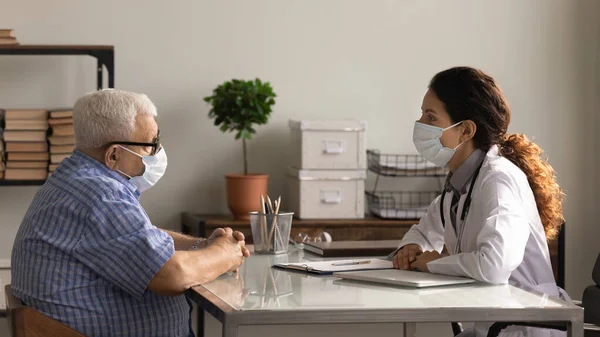 Female doctor and mature patient in facemasks consultation in hospital — Stock Photo, Image