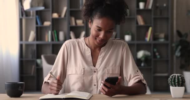 Африканская женщина сидит за столом, держа в руках смартфон, записывающий в дневник — стоковое видео