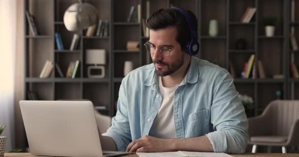 Homem focado usando fones de ouvido usando notas de escrita de laptop estudando online — Vídeo de Stock