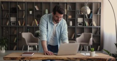 Ofiste duran adam masanın üzerine eğilip dizüstü bilgisayarla çalışıyor.