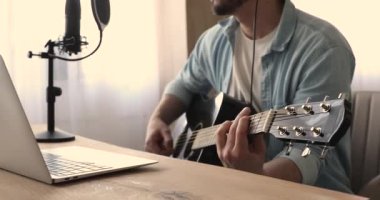 Adam gitar çalıyor. Dizüstü bilgisayarla, profesyonel mikrofonla oturuyor.