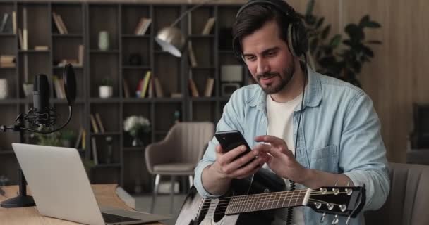 Männlicher Gitarrist sitzt mit Smartphone drinnen — Stockvideo