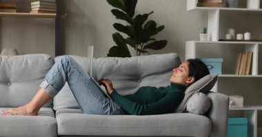 Gülümseyen genç kadın kanepede uzanmış dizüstü bilgisayarla mesaj atıyor.