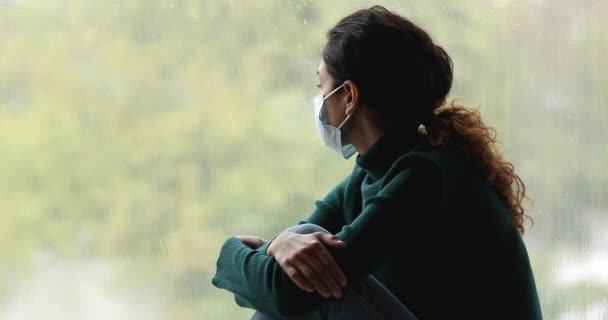 Vrouw in chirurgische gezichtsmasker kijkt uit het raam voelt zich overstuur — Stockvideo