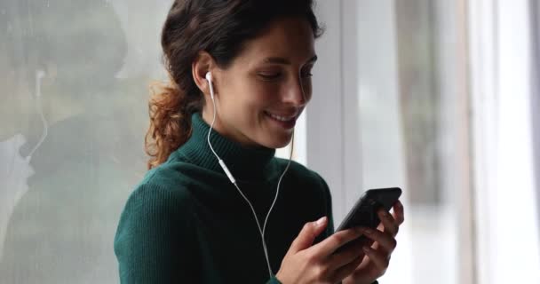Kvinne som slapper av i vinduskarmen mens hun holder smarttelefon, lytter til musikk via øretelefoner – stockvideo