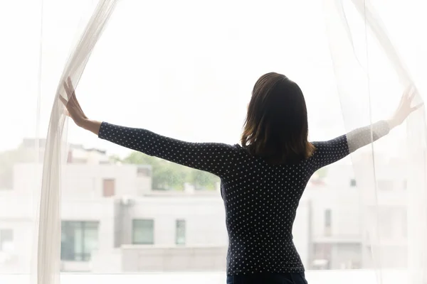 Joven mujer de raza mixta de pie cerca de cortinas de apertura ventana. — Foto de Stock