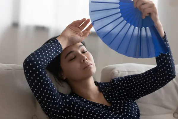 Overheated unhappy millennial asian woman using paper fan indoors. — Stock Photo, Image