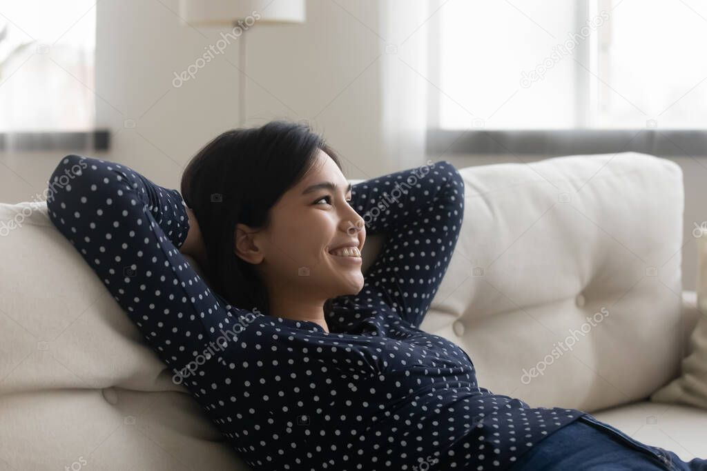 Dreamy millennial happy multiracial asian woman relaxing on cozy sofa.