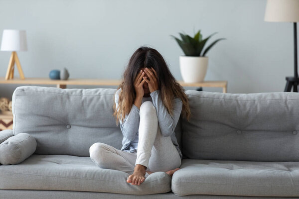 Frustrated woman covering face and crying, sitting on couch alone