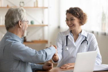 Gülümseyen genç bayan pratisyen doktor yaşlı bir hastayla el sıkışıyor..