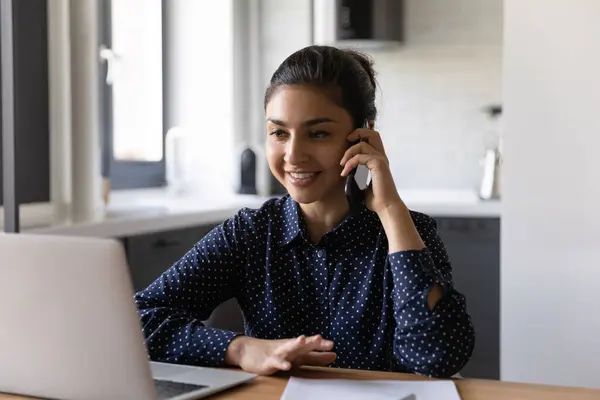 Smiling Indian woman work on laptop talking on cellphone — Stock Photo, Image