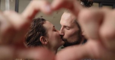 Kapalı alanda öpüşen çift parmaklarıyla kalp şekli yapıyor.