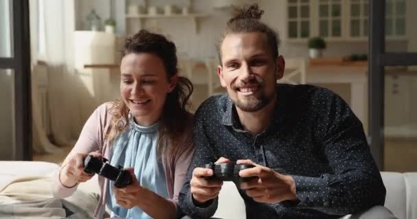 Millennial par som innehar styrenheter spelar tv-spel sittande på soffan — Stockvideo