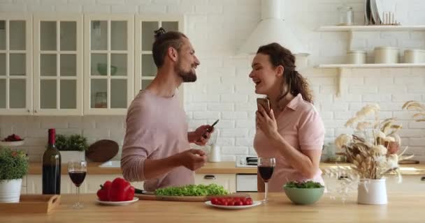 Pareja sosteniendo smartphones escuchando música bailando divirtiéndose en la cocina — Vídeos de Stock