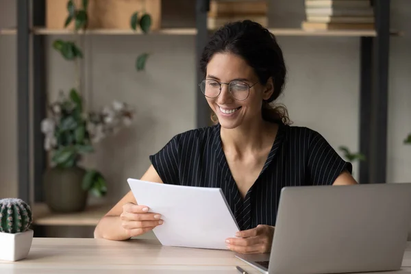 Wanita muda tersenyum membaca dokumen yang bekerja pada laptop — Stok Foto