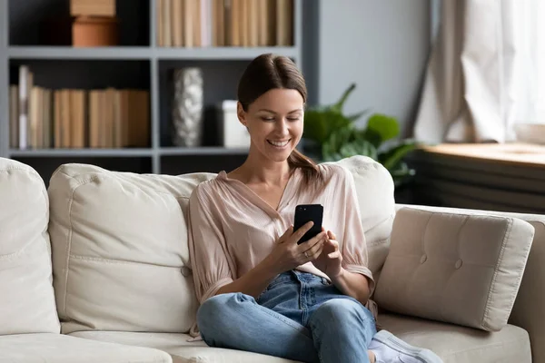 Donna sorridente che guarda lo schermo dello smartphone, seduta sul divano — Foto Stock