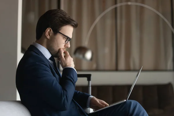Side view thoughtful businessman wearing glasses touching chin, using laptop