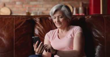 Yaşlı kadın akıllı telefonuyla evdeki rahat koltukta dinleniyor.