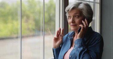 Yaşlı kadın pencerenin yanında durmuş cep telefonuyla konuşuyor.