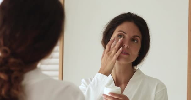 Hermosa mujer de 35 años aplicando crema hidratante, mirando en el espejo. — Vídeo de stock