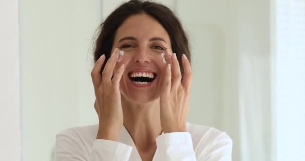 Happy laughing young woman applying cream on facial skin. — Stock Video