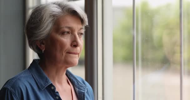 Middle-aged thoughtful female standing indoor looking out the window — Stock Video