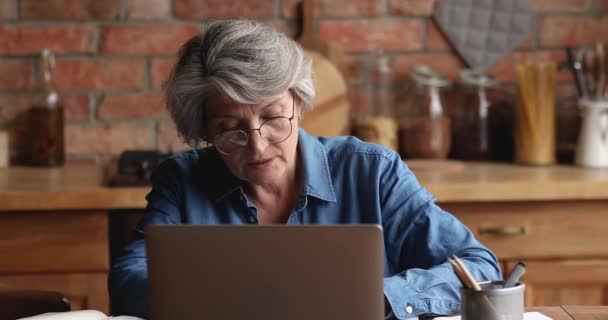 Ältere Frau sitzt drinnen und trägt Brille, die am Laptop arbeitet — Stockvideo
